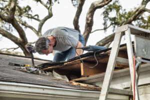 Man using crowbar to remove rotten wood from leaky roof. After removing fascia boards he has discovered that the leak has extended into the beams and decking.